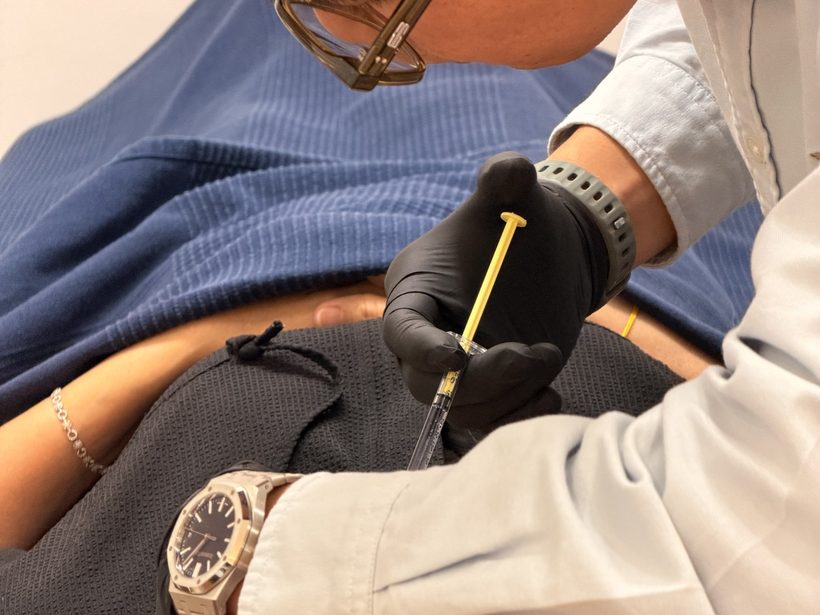 Dr Ong Jin Khang's gloved hands administering an injection into a patient's forearm in a clinic treatment room, documentary style